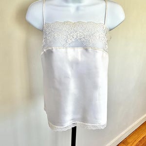 Pu&ograve; includere: Camicia di raso bianca con delicate finiture in pizzo sullo scollo e sull'orlo. La camicia presenta sottili spalline e viene esposta su un manichino bianco. Il capo ha una consistenza liscia e lucida.