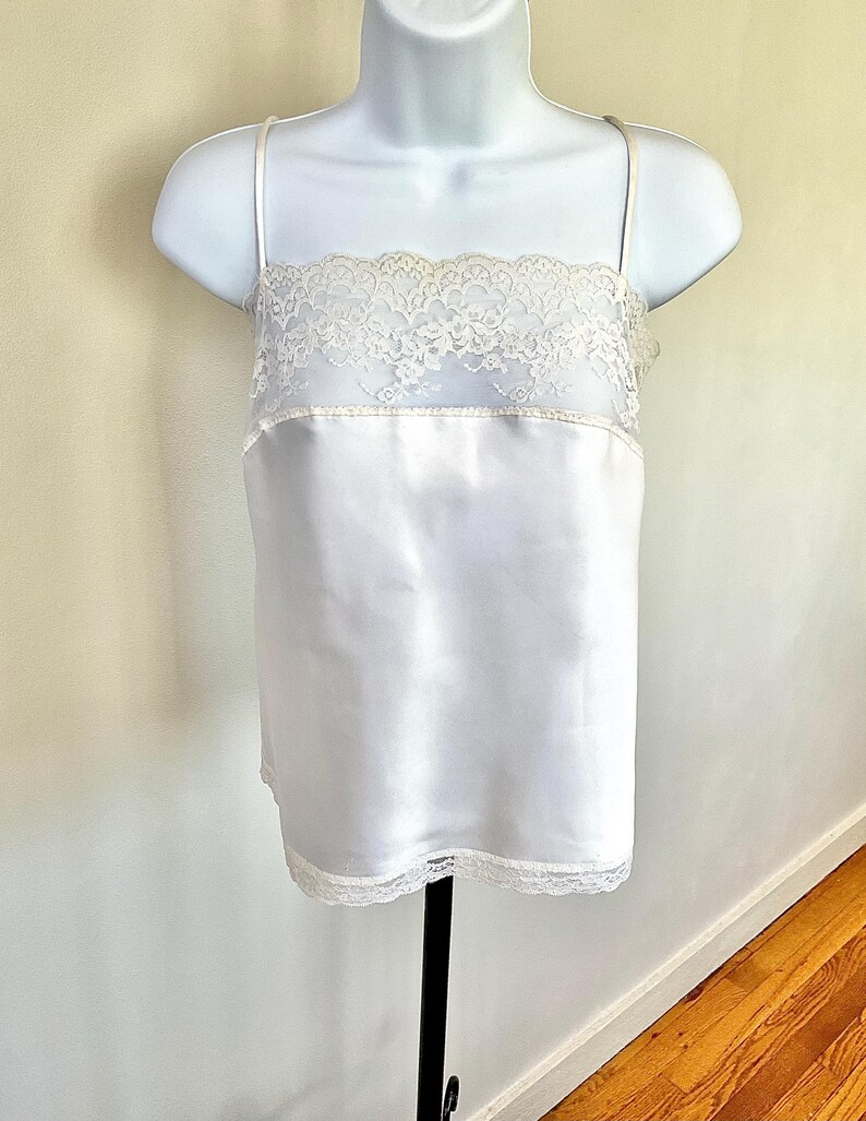 Pu&ograve; includere: Camicia di raso bianca con delicate finiture in pizzo sullo scollo e sull'orlo. La camicia presenta sottili spalline e viene esposta su un manichino bianco. Il capo ha una consistenza liscia e lucida.