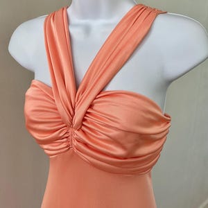 May include: A peach-colored sleeveless dress featuring a gathered, ruched bodice with a central twist detail and wide, overlapping shoulder straps that create a structured neckline on a white mannequin torso.