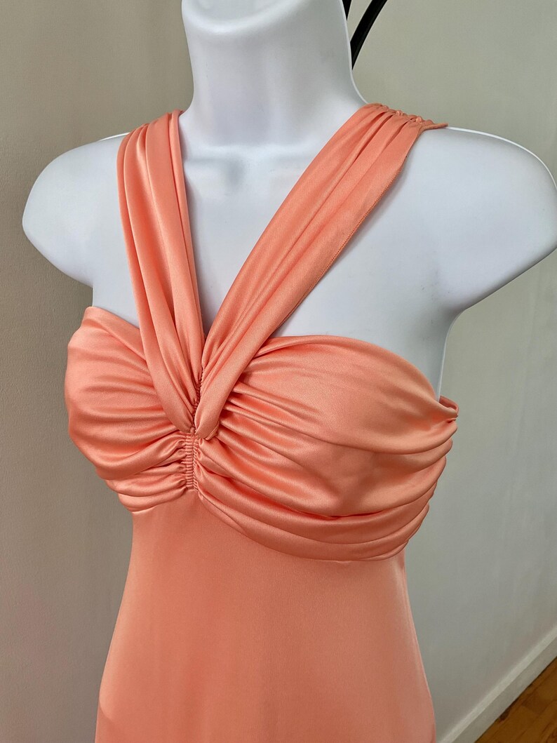 May include: A peach-colored sleeveless dress featuring a gathered, ruched bodice with a central twist detail and wide, overlapping shoulder straps that create a structured neckline on a white mannequin torso.