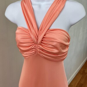 May include: A peach-colored dress with a halter neckline and a ruched bodice. The dress is displayed on a white mannequin. The fabric has a silky sheen, and the dress appears to be a formal or evening gown.