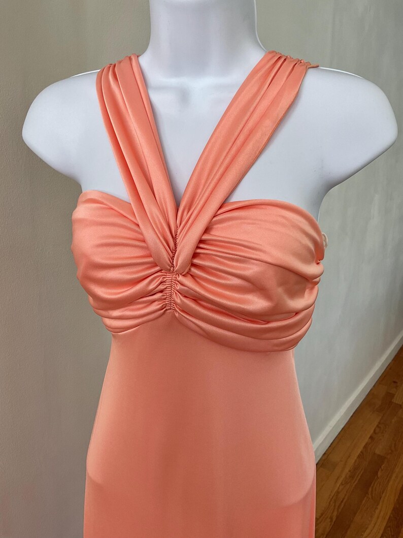 May include: A peach-colored dress with a halter neckline and a ruched bodice. The dress is displayed on a white mannequin. The fabric has a silky sheen, and the dress appears to be a formal or evening gown.