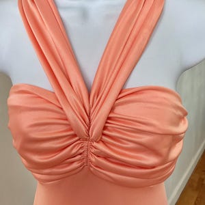 May include: A peach-colored dress with a ruched bodice and halter-style straps. The fabric has a satin sheen, and the straps cross over the chest, creating a V-neckline. The dress is displayed on a white mannequin.