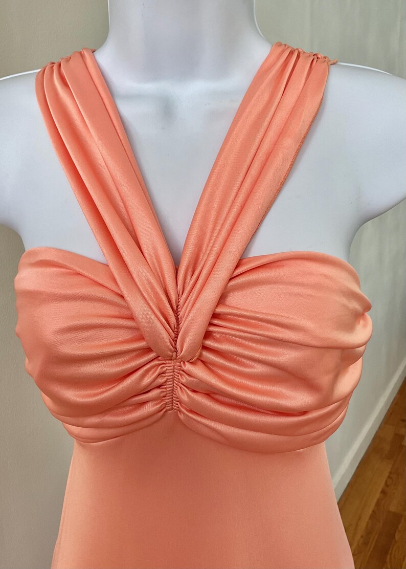 May include: A peach-colored dress with a ruched bodice and halter-style straps. The fabric has a satin sheen, and the straps cross over the chest, creating a V-neckline. The dress is displayed on a white mannequin.