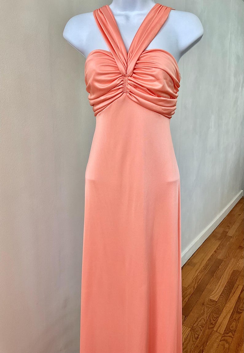 May include: A peach-colored formal gown with a halter neckline and a ruched bodice. The dress is displayed on a white mannequin and has a floor-length skirt. The fabric appears to be a soft, flowing material.
