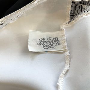 Pu&ograve; includere: Primo piano di un capo in raso bianco con delicate finiture in pizzo. Un'etichetta rettangolare con la scritta "Body Inc" &egrave; cucita sul tessuto. Il capo sembra essere lingerie vintage.