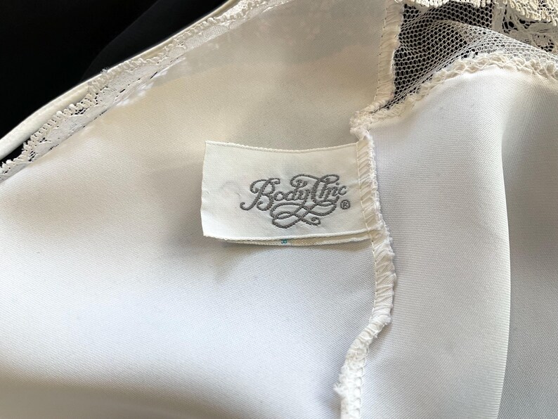 Pu&ograve; includere: Primo piano di un capo in raso bianco con delicate finiture in pizzo. Un'etichetta rettangolare con la scritta "Body Inc" &egrave; cucita sul tessuto. Il capo sembra essere lingerie vintage.