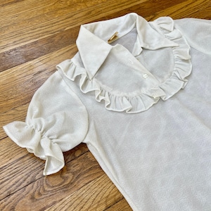 Può includere: Una blusa bianca a maniche corte con colletto e polsini a balze. La blusa ha una chiusura con bottone e un tessuto strutturato.