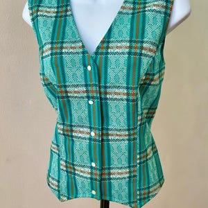 May include: A sleeveless vest featuring a teal, white, and brown plaid pattern with a V-neckline and a front button closure. The fabric displays a repeating geometric texture within the plaid design, and the garment is displayed on a mannequin.