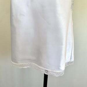 Pu&ograve; includere: Un sottoveste in raso bianco con un delicato bordo in pizzo sull'orlo. L'abito &egrave; esposto su un supporto nero. Il tessuto ha una lucentezza sottile.
