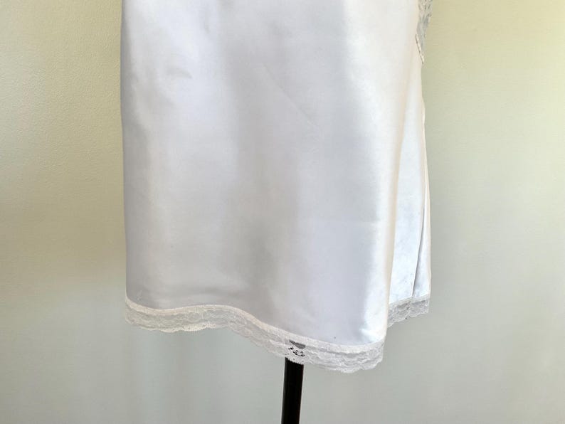 Pu&ograve; includere: Un sottoveste in raso bianco con un delicato bordo in pizzo sull'orlo. L'abito &egrave; esposto su un supporto nero. Il tessuto ha una lucentezza sottile.