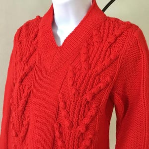 Puede incluir: Suéter de punto rojo con un patrón de punto de cable y cuello en V.