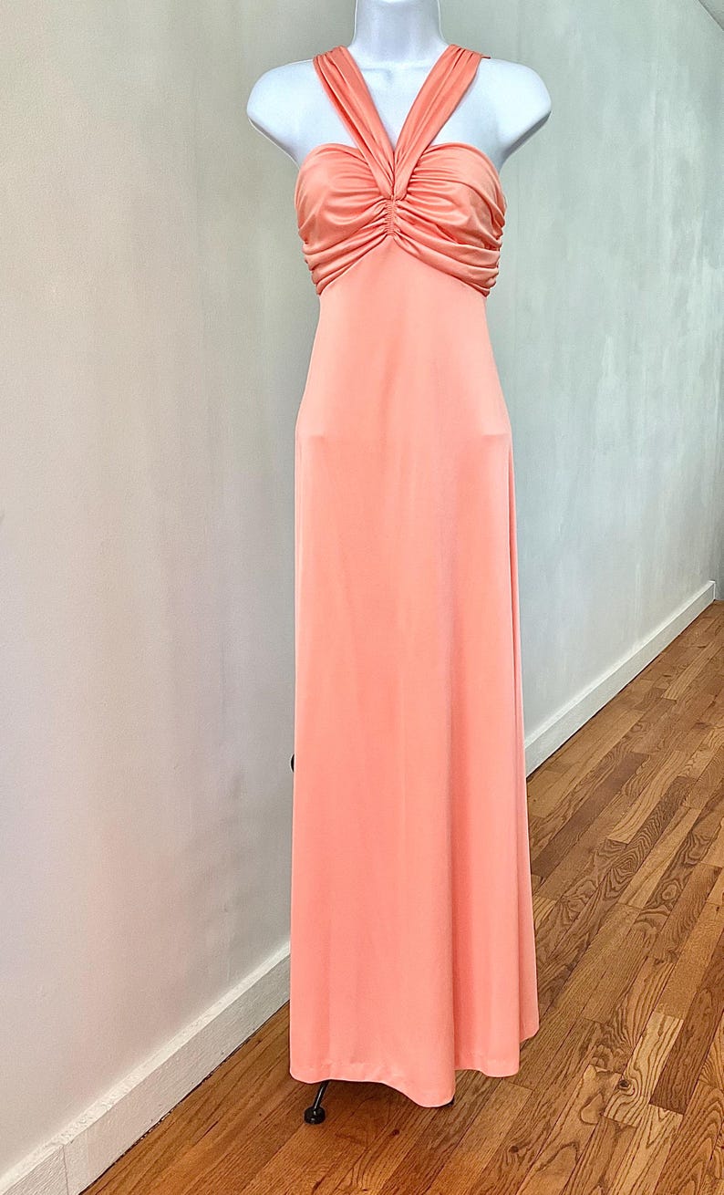 May include: A floor-length peach-colored gown featuring a halter neckline with a twisted, ruched bodice detail. The dress has a smooth, solid-colored fabric that flows from the fitted waist down to the hem, creating a clean and simple silhouette.