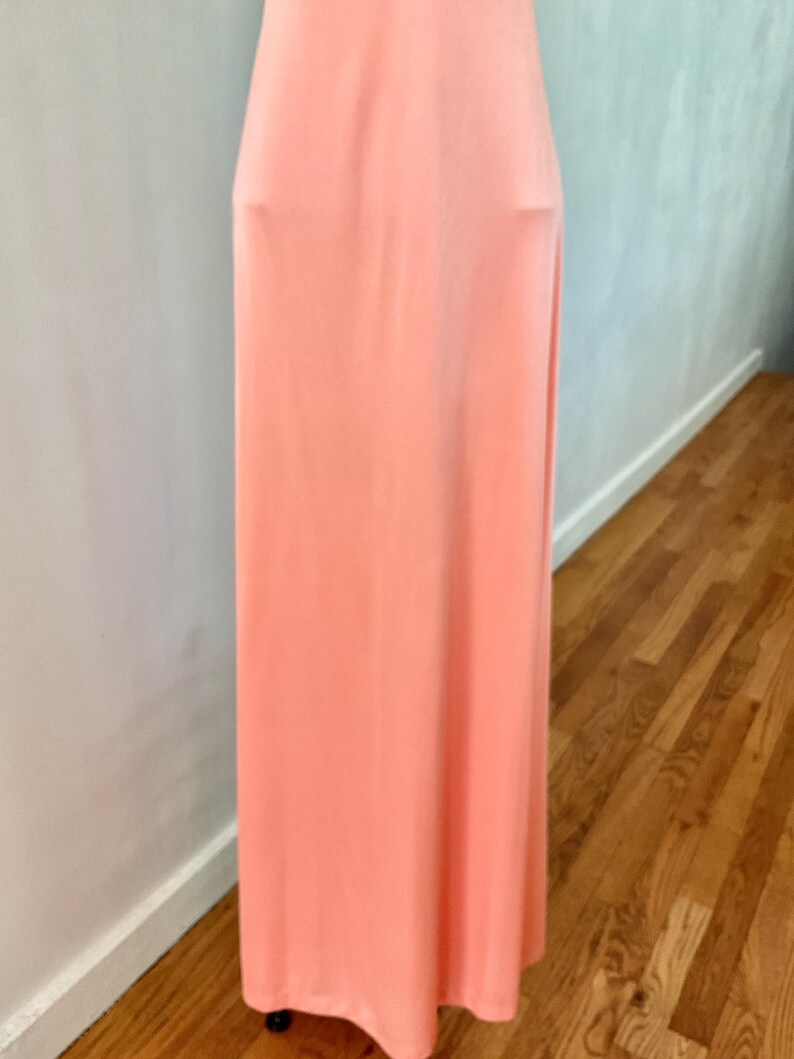 May include: A floor-length, peach-colored dress. The dress has a simple, flowing design with a high waistline. The fabric appears to be a soft, lightweight material. The dress is displayed against a neutral background.