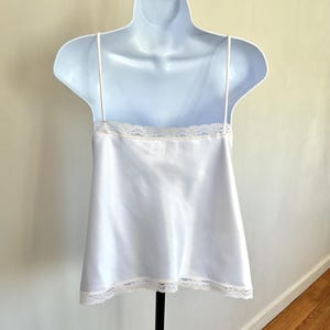 Pu&ograve; includere: Una camisole di raso bianco con delicate spalline sottili e finiture in pizzo lungo lo scollo e l'orlo. La camisole &egrave; esposta su un manichino bianco, che mette in risalto il suo design semplice ma elegante. Il capo &egrave; un classico della lingerie.