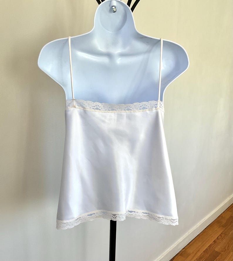 Pu&ograve; includere: Una camisole di raso bianco con delicate spalline sottili e finiture in pizzo lungo lo scollo e l'orlo. La camisole &egrave; esposta su un manichino bianco, che mette in risalto il suo design semplice ma elegante. Il capo &egrave; un classico della lingerie.