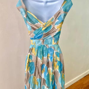 Puede incluir: Vestido con estampado floral sobre un maniqu&iacute;. El vestido presenta un escote en V y una cintura entallada. La tela tiene un estampado de flores azules, amarillas y blancas sobre un fondo topo. El vestido es sin mangas con una falda plisada.