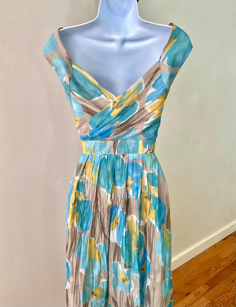 Puede incluir: Vestido con estampado floral sobre un maniqu&iacute;. El vestido presenta un escote en V y una cintura entallada. La tela tiene un estampado de flores azules, amarillas y blancas sobre un fondo topo. El vestido es sin mangas con una falda plisada.
