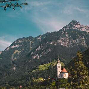 May include: Scenic landscape featuring a white church with a clock tower and a red roof nestled in a valley. Towering mountains covered in lush green trees dominate the background under a clear blue sky. A road and foliage are in the foreground.