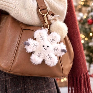 May include: A plush white snowflake keychain with a smiling face, embroidered gray details, and a small beige pom-pom attached to a gold-toned metal clasp. The snowflake is made of soft, fuzzy material and hangs from a brown leather handbag.