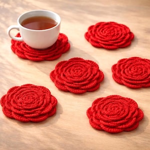May include: A white teacup sits on a red crocheted coaster shaped like a rose. Several other red rose-shaped coasters are scattered around. The coasters are made of a textured yarn and are on a light brown surface.