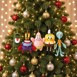 Op de afbeelding: Vijf gehaakte ornamenten die stripfiguren voorstellen, hangend in een kerstboom. De figuren bestaan uit een groene cilinder, een rode krab, een roze zeester, een geel vierkant en een lichtblauwe koppotige, allemaal aan gouden koordjes.