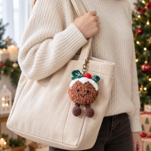 May include: A plush Christmas pudding keyring featuring a brown textured body, a white icing top with a red pom-pom berry, and two green leaves. The round character has black embroidered eyes and a small smile, with two dangling brown legs.