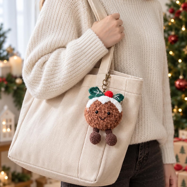 May include: A plush Christmas pudding keyring featuring a brown textured body, a white icing top with a red pom-pom berry, and two green leaves. The round character has black embroidered eyes and a small smile, with two dangling brown legs.