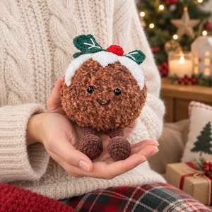 May include: A brown crocheted Christmas pudding plush toy with a white frosting top, green leaves, and a small red berry. The plush features black safety eyes, a stitched smile, and two small dangling brown crocheted legs with round feet.