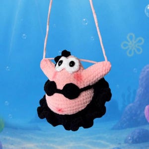 May include: A pink crocheted bag shaped like a starfish character with large white eyes, black pupils, and a black bikini top. The bag features a ruffled black skirt at the base and includes a thin pink crocheted strap for carrying over the shoulder.