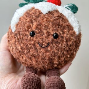 May include: A round, brown, fuzzy crochet plush toy shaped like a Christmas pudding. It features a white textured top, a small red berry, and two green leaves. The face has two black bead eyes and a stitched black smile, with two dangling brown feet.