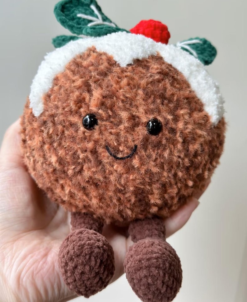 May include: A round, brown, fuzzy crochet plush toy shaped like a Christmas pudding. It features a white textured top, a small red berry, and two green leaves. The face has two black bead eyes and a stitched black smile, with two dangling brown feet.