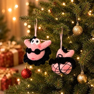 May include: Two handmade crochet ornaments shaped like pink starfish characters. Each figure features large white eyes, a black top, and a black skirt. The ornaments are suspended by light pink yarn loops, showcasing detailed stitching and textures.