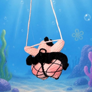 May include: A light pink crocheted starfish plush toy wearing a black tutu and black fishnet stockings. The plush has a small black hat on its head and is suspended by thin light pink strings against a blue underwater background.