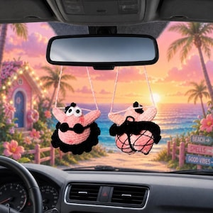 May include: Two crocheted pink starfish figures hanging from a rearview mirror. Each figure features large white eyes, a black top, and a black ruffled skirt. The figures are suspended by thin pink strings against a tropical beach background.