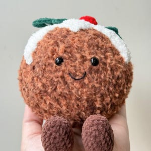May include: A round, brown, fuzzy plush toy shaped like a Christmas pudding. It features a white textured top layer, a small red berry accent, and two dark green leaves. The face has two black bead eyes and a stitched black smile.