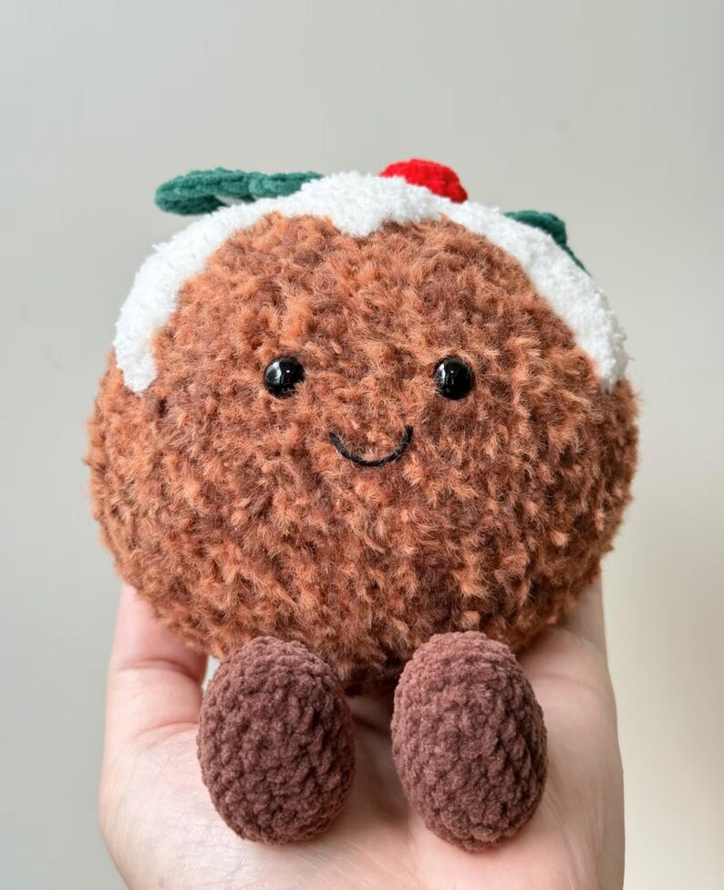 May include: A round, brown, fuzzy plush toy shaped like a Christmas pudding. It features a white textured top layer, a small red berry accent, and two dark green leaves. The face has two black bead eyes and a stitched black smile.