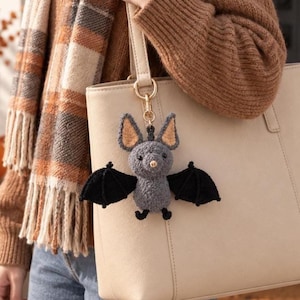May include: A gray crocheted bat keychain with black wings and tan inner ears, attached to a gold-toned metal clasp. The bat has a textured, fuzzy appearance, small black eyes, and a tiny nose, hanging from a beige leather-like tote bag.