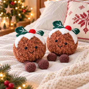 May include: Two round, brown, fuzzy crochet plushies shaped like Christmas puddings. Each features a white icing top, a small red berry, and two green leaves. They have black bead eyes, embroidered smiles, and dangling brown feet.