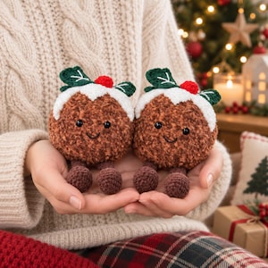 May include: Two brown crocheted plush toys shaped like Christmas puddings, each featuring a white frosting top, green leaves, a small red berry, black bead eyes, a stitched smile, and two dangling brown crocheted legs with round feet.