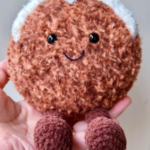 May include: A round, brown, fuzzy plush toy shaped like a Christmas pudding. It features a white crocheted topping, a small red berry, and green leaves on top. The face has two black plastic eyes and a stitched black smile, with two small brown legs.