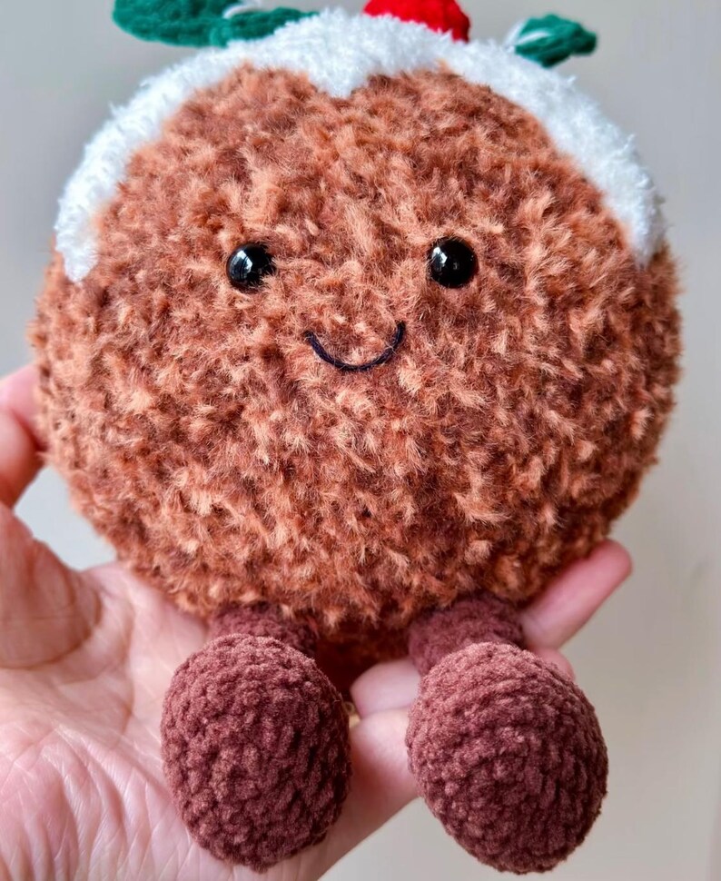 May include: A round, brown, fuzzy plush toy shaped like a Christmas pudding. It features a white crocheted topping, a small red berry, and green leaves on top. The face has two black plastic eyes and a stitched black smile, with two small brown legs.