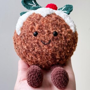 May include: A round, brown, fuzzy crochet plush toy shaped like a Christmas pudding. It features a white textured top, two dark green leaves, a small red berry, black bead eyes, and a stitched smile, with two small brown dangling feet.