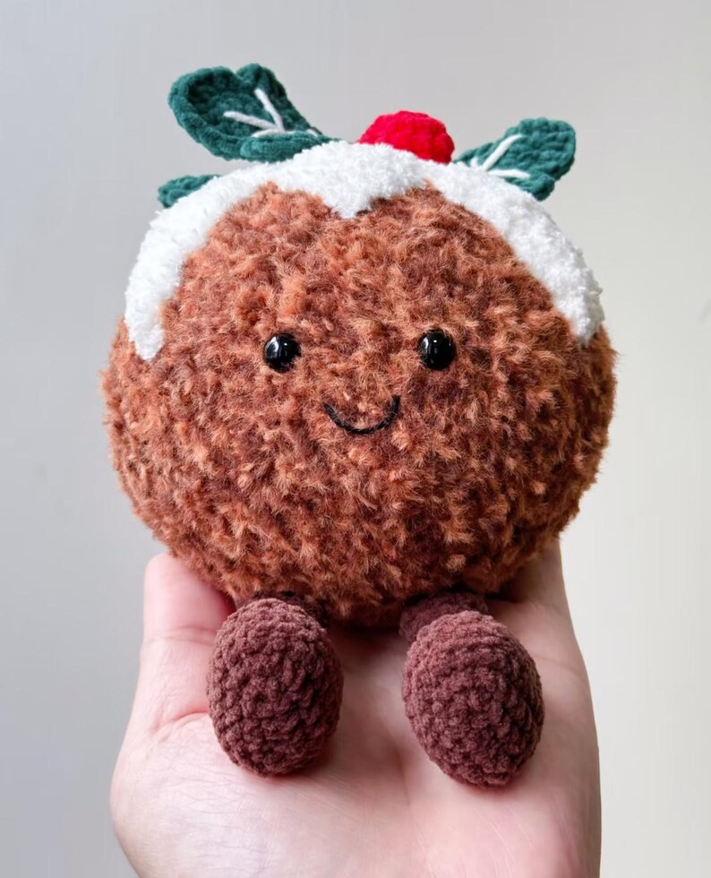 May include: A round, brown, fuzzy crochet plush toy shaped like a Christmas pudding. It features a white textured top, two dark green leaves, a small red berry, black bead eyes, and a stitched smile, with two small brown dangling feet.