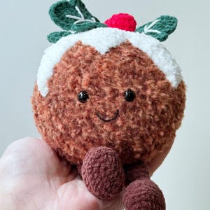 May include: A round, brown, fuzzy crochet plush toy shaped like a Christmas pudding. It features a white textured top, two dark green leaves with white stitching, a small red berry, black bead eyes, and a stitched smile with two dangling legs.