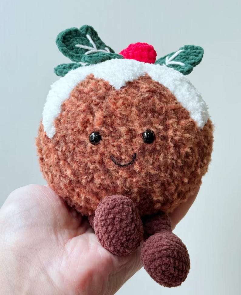 May include: A round, brown, fuzzy crochet plush toy shaped like a Christmas pudding. It features a white textured top, two dark green leaves with white stitching, a small red berry, black bead eyes, and a stitched smile with two dangling legs.
