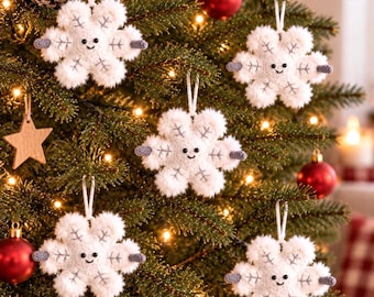 May include: Five white plush snowflake ornaments with fuzzy textures, each featuring a smiling face with two black bead eyes, a small embroidered mouth, and grey stitched details on the points, hanging from white strings on a green tree.