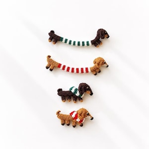 Puede incluir: Cuatro figuras de perro salchicha tejidas a ganchillo. Dos son alargadas con cuerpos a rayas en verde-blanco y rojo-blanco. Dos son más pequeñas, de pie, con bufandas tejidas en verde-blanco y rojo-blanco. Todas tienen hilo marrón y detalles negros.