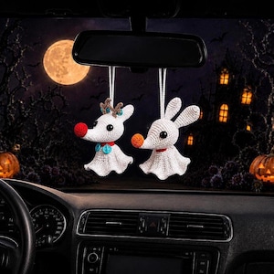 May include: Two white crocheted ghost-like dog ornaments hanging from a rearview mirror. One figure has a red nose, brown antlers, and blue buttons, while the other has an orange nose and long ears. Both have black eyes and red collars.