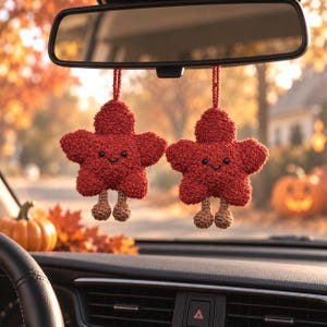 Pode incluir: Dois ornamentos suspensos para carro em forma de estrelas de cinco pontas feitas de fio vermelho texturizado. Cada estrela apresenta um pequeno rosto sorridente preto bordado e duas pernas castanhas pendentes com pés redondos, suspensas no retrovisor.
